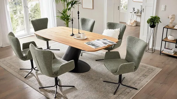 Esszimmer mit großem Holztisch und sechs grünen Stühlen auf einem Teppich. Moderne Einrichtungsgegenstände und Pflanzen ergänzen das Ambiente.