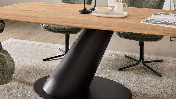 Moderner Esstisch mit hölzerner Tischplatte und schwarzem, schrägem Metallfuß, umgeben von grünen Stühlen auf Teppich.