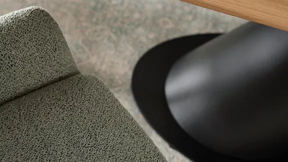 Detailaufnahme von einer grau-grünen Textilstuhloberfläche neben einem Holzdesktisch und schwarzem Tischfuß auf gemustertem Teppich.
